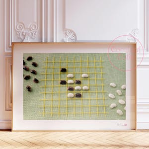 Puede incluir: Una impresión enmarcada de un juego de Go con frijoles negros y blancos dispuestos en un patrón sobre un fondo verde. El tablero está hecho de cuerda amarilla.