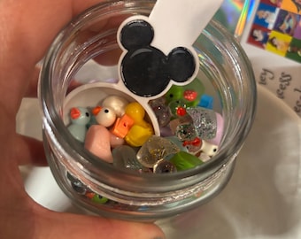 Puede incluir: Un tarro de cristal transparente lleno de figuras en miniatura de colores, incluyendo patos, ratones y otros objetos pequeños. Una etiqueta blanca con una cabeza de Mickey Mouse negra está colocada encima del tarro.