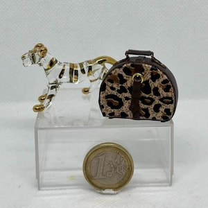 May include: A clear glass figurine of a tiger with gold accents and stripes, next to a small brown and tan leopard print handbag. A one Euro coin is in the foreground.