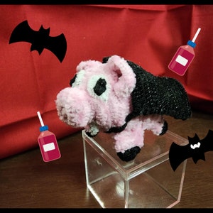 May include: A pink plush pig wearing a black cape and fangs, standing on a clear cube with two black bat silhouettes in the background.