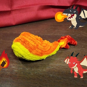 May include: A yellow and orange pipe cleaner whale with a red and orange pipe cleaner tail. The whale is on a wooden surface with two cartoon dragons, one black and one red, in the background.