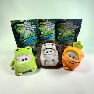 Op de afbeelding: Drie pluche speeltjes in de vorm van een kikker, een egel en een wortel, elk met een klein wit gezichtje zichtbaar door een opening. Daarachter staan drie identieke donkergroene productverpakkingen met de tekst "Mutmacher monster" en "Welches Monster hat sich hier versteckt?" op de voorzijde.
