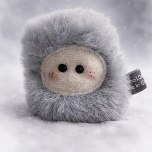 Könnte beinhalten: Ein kuscheliges, graues, pelziges Wesen mit einem hellbeigen Gesicht, schwarzen Knopfaugen und rosigen Wangen. Das Spielzeug hat ein Etikett an der Seite. Der Hintergrund ist eine weiche, verschwommene Schneelandschaft.