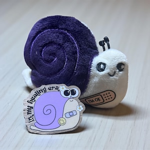 Könnte beinhalten: Ein Plüsch-Schnecken-Spielzeug mit einem lila Gehäuse und weißem Körper, neben einer Holz-Anstecknadel. Die Anstecknadel zeigt eine Cartoon-Schnecke mit einem Pflaster und dem Text "in my healing era". Das Plüschtier hat ein Pflaster mit der Aufschrift "I'm ok".