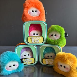 Könnte beinhalten: Eine Sammlung farbenfroher Plüschmonster-Spielzeuge, jedes mit einem flauschigen Äußeren und einem niedlichen, runden Gesicht mit zwei schwarzen Augen und rosigen Wangen. Die Spielzeuge befinden sich in quadratischen Boxen mit dem Text "Monster Box". Farben: Orange, Grün, Blau, Gelb und Lila.