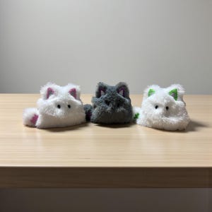 Könnte beinhalten: Drei Plüschkatzenspielzeuge in einer Reihe. Die Katzen sind weiß, grau und weiß. Die weißen Katzen haben rosa Akzente an Ohren und Pfoten, während die graue Katze lila Akzente hat. Die Katzen liegen auf einer Holzoberfläche.
