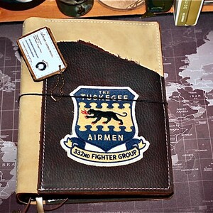 May include: A leather journal with a tan and dark brown cover. The front features a patch with the words "THE TUSKEGEE AIRMEN 332ND FIGHTER GROUP" and a black panther emblem. A small tag is attached to the journal.