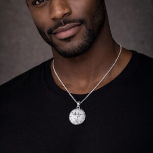 A man wearing a black t-shirt and a silver necklace with a pendant, looking directly at the camera with a serious expression.