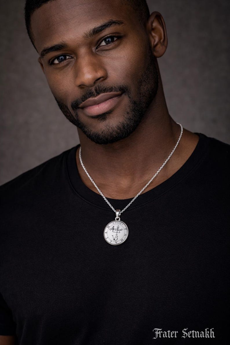 A man wearing a black t-shirt and a silver necklace with a pendant, looking directly at the camera with a serious expression.