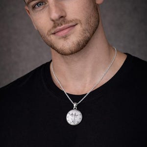 A man wearing a black t-shirt and a silver necklace with a pendant is shown in the image.
