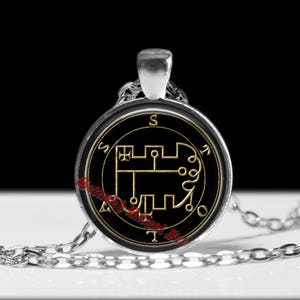 Puede incluir: Un collar con colgante circular de tono plateado que presenta un fondo negro con un diseño de sello dorado. Las letras S, T, A y T están colocadas alrededor del perímetro del emblema circular. El texto Setnak Ritual Art está en rojo.