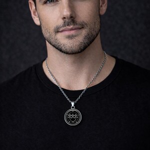 A man wearing a black shirt and a silver necklace with a pendant, looking directly at the camera.
