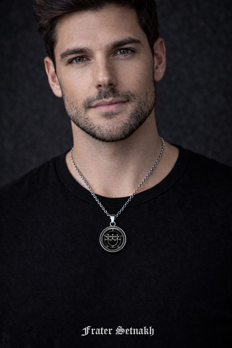 A man wearing a black shirt and a silver necklace with a pendant, looking directly at the camera.