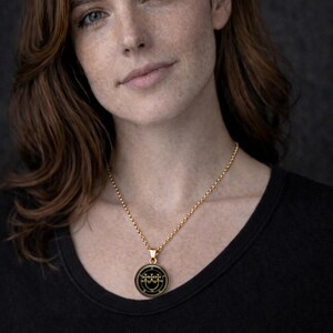 A portrait of a woman with long brown hair wearing a black top and a gold necklace with a pendant.