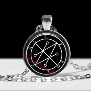 May include: A silver pendant necklace with a circular black charm displaying a white geometric design. The pendant is suspended from a fine silver chain. The design incorporates lines, circles, and symbols.