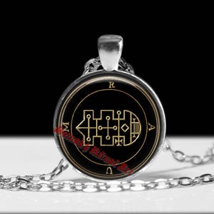 May include: A silver-toned pendant necklace featuring a circular glass cabochon. The black insert displays a gold-coloured geometric sigil with the letters R, A, and M positioned around the perimeter. The text Setnakh Ritual Art is printed in red.