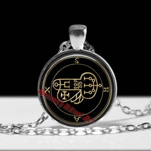 Op de afbeelding: Een zilverkleurige ronde hanger aan een ketting met een zwarte inzet en een goudkleurig geometrisch zegelontwerp. De hanger hangt aan een bijpassende zilverkleurige metalen ketting. De tekst "Seinakh Ritual Art" is in het rood op het midden gedrukt.