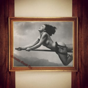 May include: A black and white photograph of a woman flying on a broomstick. The woman is wearing a black garter belt and is looking up at the sky. The photograph is framed in a wooden frame.