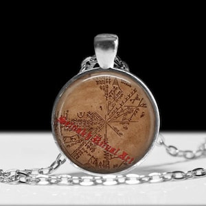 May include: A silver-toned pendant necklace featuring a circular pendant with a map design in shades of brown and red. The pendant is suspended from a silver chain. The text "Sacred Ritual Art" is printed on the pendant.