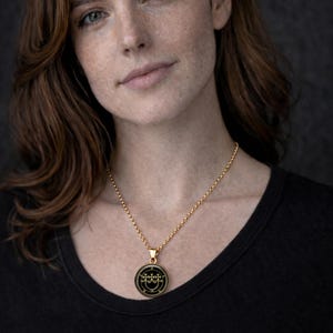A portrait of a woman with long brown hair wearing a black top and a gold necklace with a pendant.