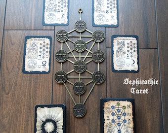 May include: A Sephirothic Tree of Life metal wall hanging with a keyhole-shaped top, surrounded by tarot cards. The cards and the tree are on a wooden surface. The text "Sephirothic Tarot" is visible.