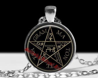 May include: A silver-toned pendant necklace featuring a black circular charm with a gold pentagram design and inscribed text. The pendant hangs from a silver chain, set against a white surface.