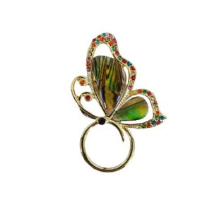 May include: A gold-coloured butterfly ring featuring iridescent abalone shell wings and a ring base. The butterfly's edges are decorated with colourful rhinestones. The ring is set against a white background.