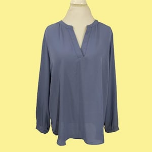 Puede incluir: Una blusa de manga larga de color azul bígaro con cuello en V. La blusa se exhibe en un maniquí sobre un fondo amarillo. Las mangas son largas y terminan en un puño en la muñeca.