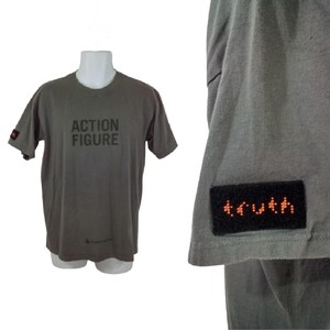 Könnte beinhalten: Ein graues T-Shirt mit dem Aufdruck "ACTION FIGURE" auf der Vorderseite. Das Shirt hat einen schwarzen Patch auf dem Ärmel mit dem Wort "truth" in Orange.