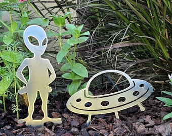 Puede incluir: Una silueta de metal de un extraterrestre de pie junto a una silueta de metal de un platillo volante. Ambos están colocados en un lecho de mantillo marrón.