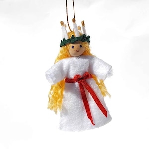 May include: A white felt doll with blonde hair wearing a green wreath with white candles and a red ribbon. The doll has a wooden head with a painted face.
