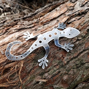 May include: A silver metal gecko with a spotted pattern, sitting on a piece of weathered wood.