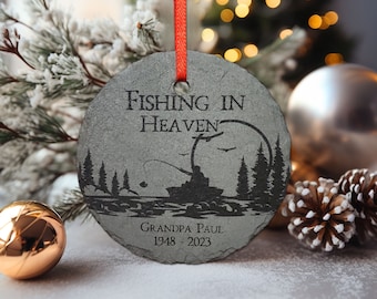 May include: A gray slate Christmas ornament with a black silhouette of a person fishing in a lake with trees in the background. The ornament reads "Fishing in Heaven" and "Grandpa Paul 1948 - 2023".