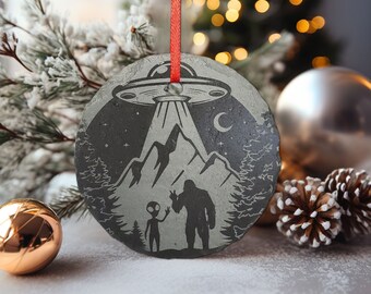 May include: A black slate Christmas ornament with a gray alien and a Bigfoot standing in front of a mountain range with a UFO above them. The ornament has a red ribbon attached to it.