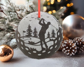 Può includere: Ornamento natalizio rotondo a tema golf con un nastro rosso. L'ornamento presenta una silhouette di un golfista che fa swing, alberi e una buca da golf. L'ornamento è grigio e ha una superficie strutturata. Una palla d'oro e d'argento e delle pigne sono sullo sfondo.