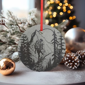 May include: A round, gray slate ornament with a black and white illustration of a hiker with a backpack walking through a mountain landscape. The ornament has a red ribbon attached for hanging.