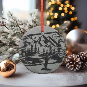 May include: A round, gray slate Christmas ornament with a detailed etched design of a mountain landscape, a person fishing, and trees. The ornament is suspended by a red ribbon. A gold ornament and pinecones are in the foreground.