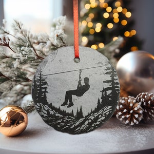 May include: A round, gray slate ornament with a silhouette of a person ziplining through a forest. The ornament is tied with a red ribbon.