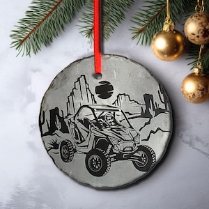 May include: A round, gray slate Christmas ornament with a black and white silhouette of a person driving a dune buggy in a desert landscape. The ornament has a red ribbon attached to the top.
