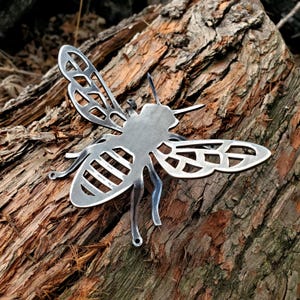 May include: A silver metal bee sculpture with intricate wing cutouts. The bee is resting on a textured, brown tree trunk. The sculpture has detailed antennae and legs, and is designed to be a decorative garden accent.