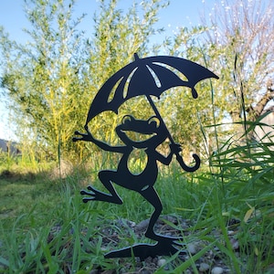 May include: A black metal frog garden stake holding an umbrella. The frog is depicted with a smiling face and outstretched arms, standing on one leg. The umbrella has a curved handle and is open, with a decorative design.