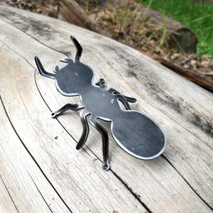 May include: A black metal ant silhouette, with a detailed design, is displayed on a wooden surface. The ant is facing upwards, with its legs and antennae visible.