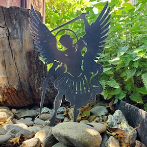 K&ouml;nnte beinhalten: Ein schwarzer Metall-Gartenstecker in Form einer Silhouette, die einen Engel mit gro&szlig;en Fl&uuml;geln, einem Heiligenschein und einem erhobenen Schwert darstellt. Die Figur ist flach ausgeschnitten mit detaillierten Federn und flie&szlig;endem Gewand.