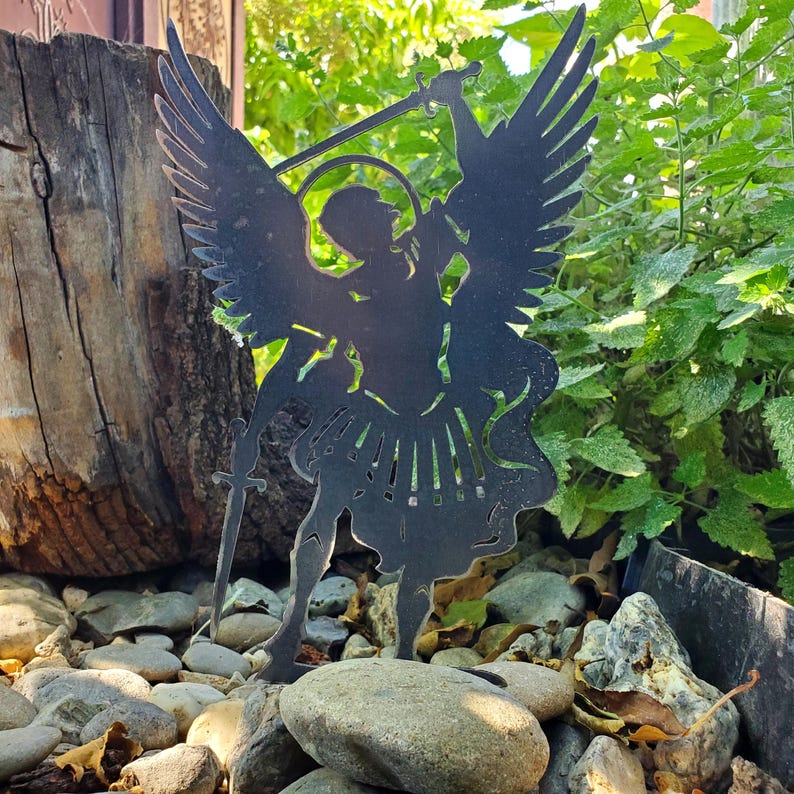 K&ouml;nnte beinhalten: Ein schwarzer Metall-Gartenstecker in Form einer Silhouette, die einen Engel mit gro&szlig;en Fl&uuml;geln, einem Heiligenschein und einem erhobenen Schwert darstellt. Die Figur ist flach ausgeschnitten mit detaillierten Federn und flie&szlig;endem Gewand.