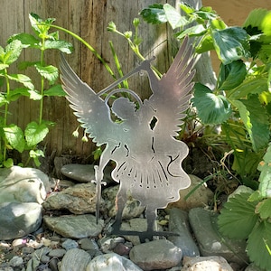 K&ouml;nnte beinhalten: Ein flacher, metallischer, silberfarbener Gartenstecker mit der ausgeschnittenen Silhouette eines Engels mit gro&szlig;en, ausgebreiteten Fl&uuml;geln, einem Heiligenschein und einem in einer Hand gehaltenen Schwert. Die Figur zeigt eine dynamische Pose.