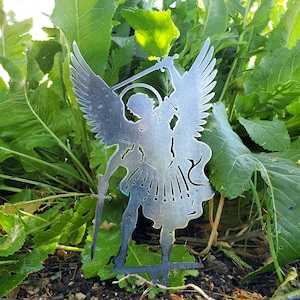 K&ouml;nnte beinhalten: Eine silberfarbene Metallsilhouette einer Engelsfigur mit gro&szlig;en Fl&uuml;geln, einem Heiligenschein und einem aufrecht gehaltenen Schwert. Das ausgeschnittene Design zeigt detaillierte Formen, einschlie&szlig;lich flie&szlig;ender Gew&auml;nder und definierter Federn.