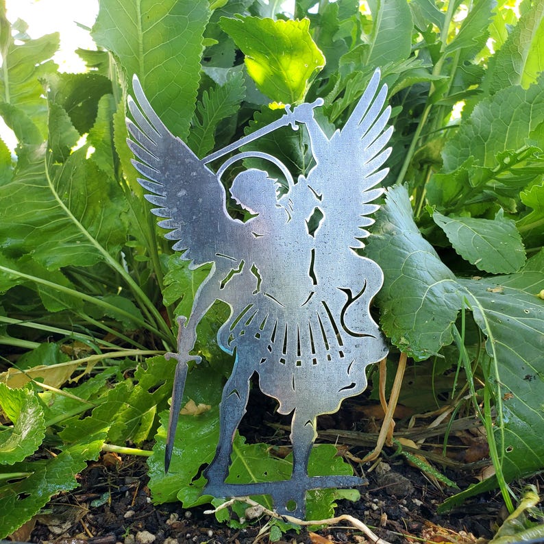K&ouml;nnte beinhalten: Eine silberfarbene Metallsilhouette einer Engelsfigur mit gro&szlig;en Fl&uuml;geln, einem Heiligenschein und einem aufrecht gehaltenen Schwert. Das ausgeschnittene Design zeigt detaillierte Formen, einschlie&szlig;lich flie&szlig;ender Gew&auml;nder und definierter Federn.