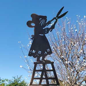 May include: A metal garden art piece depicts a mouse figure holding scissors, standing on a small ladder. The mouse wears a dress with floral cutouts. The silhouette is against a bright blue sky and a tree with white blossoms.