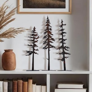 May include: Three metal tree sculptures in varying heights, with a rustic brown finish. The trees are displayed on a white shelf, alongside a vase of dried plants and a framed landscape print. The decor adds a natural, outdoorsy touch to the space.