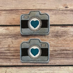 May include: Two gray felt camera patches with black details and a teal heart in the center of the lens. The lens is surrounded by silver stitching. The patches are on a wooden background.